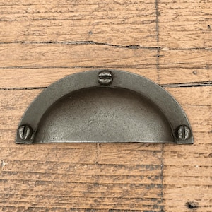 May include: A semi-circular metal drawer pull with a dark, weathered finish. The handle features a recessed cup design and is secured by three visible flat-head screws, one at the top centre and two at the bottom corners of the curved frame.