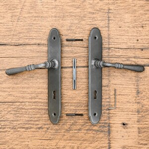 Può includere: Un set di due maniglie per porta in metallo grigio scuro con placche rettangolari lunghe, ciascuna dotata di foro per chiave. Il set include un perno quadro centrale, due viti di montaggio orizzontali e una piccola chiave a brugola.