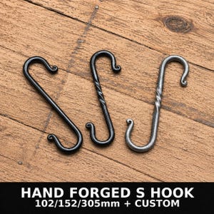 Può includere: Tre ganci a S in metallo con estremità arricciate. Due ganci sono nero opaco e uno è color argento metallizzato. I due ganci a destra presentano un design centrale ritorto. Il testo in basso recita: HAND FORGED S HOOK 102/152/305mm + CUSTOM.