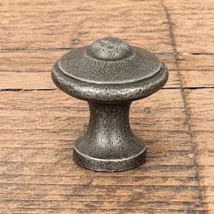 Könnte beinhalten: Ein runder Möbelknopf mit einer verwitterten, dunkelgrauen Metalloberfläche. Der Knopf verfügt über eine breite, flache Oberseite mit einer zentralen, gewölbten Erhebung und einen konischen, zylindrischen Sockel, der sich nach unten hin verbreitert.