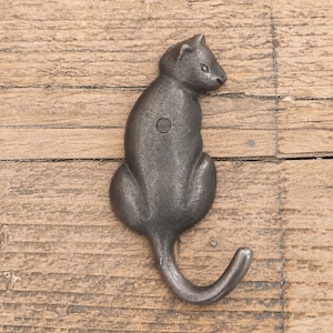 May include: A dark grey, cast metal wall hook shaped like a sitting cat. The cat is depicted in profile, facing right, with its tail curving upwards to form the hook. A single screw head is visible in the centre of the cat's body for mounting.