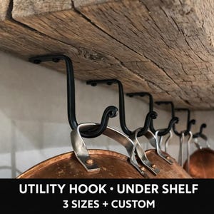 Könnte beinhalten: Schwarze Metall-Haken unter einem Holzregal, die Kupfer-Töpfe und -Pfannen halten. Die Haken haben ein dekoratives Scroll-Design. Der Text unten lautet: UTILITY HOOK • UNDER SHELF 3 SIZES + CUSTOM.