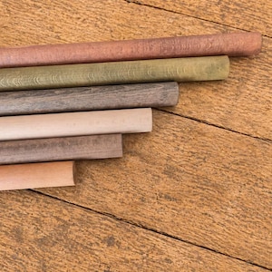 May include: A collection of wooden dowels in various colors, including shades of brown, green, and beige, arranged diagonally on a wooden surface. The dowels are cylindrical and appear to be new.
