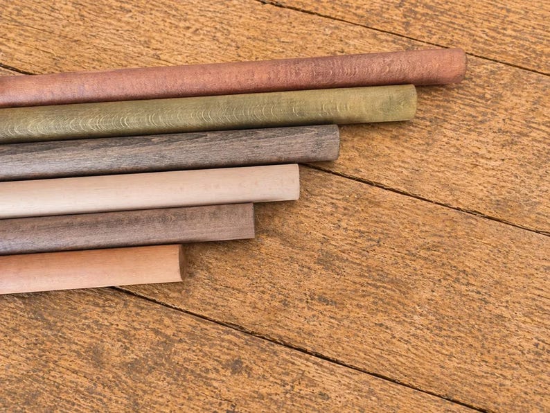 May include: A collection of wooden dowels in various colors, including shades of brown, green, and beige, arranged diagonally on a wooden surface. The dowels are cylindrical and appear to be new.