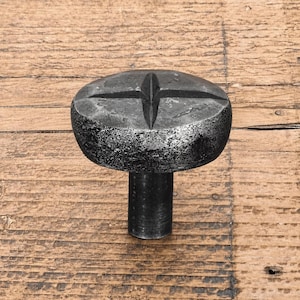 Puede incluir: Un pomo de mueble de metal gris oscuro con una superficie superior redondeada y texturizada que presenta un diseño prominente y elevado de estrella o cruz de cuatro puntas. El pomo tiene una base cilíndrica y un acabado mate rugoso.