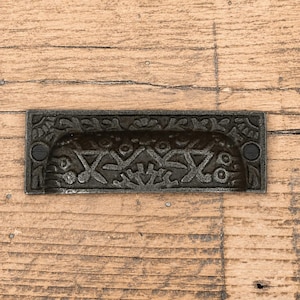 May include: A rectangular dark grey metal bin pull handle featuring a recessed grip area and an embossed floral and geometric pattern across the front surface. The hardware includes two black screws positioned on the left and right sides.