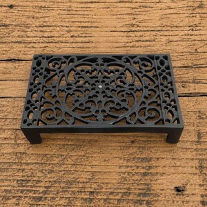 Puede incluir: Un salvamanteles rectangular de metal negro con un intrincado diseño calado que presenta un motivo circular central rodeado de patrones de volutas. La pieza se apoya sobre cuatro patas cortas y resistentes en cada esquina.