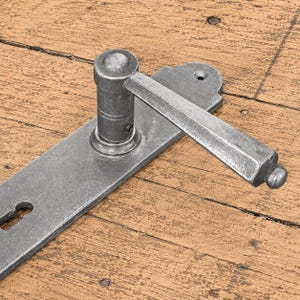 Puede incluir: Un juego de manilla de puerta de metal plateado con una placa rectangular larga que tiene una abertura para llave en un extremo y una manilla de palanca unida a una base cilíndrica. Los herrajes tienen un acabado mate texturizado.