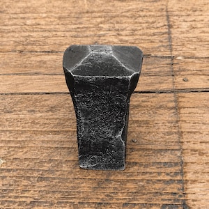 May include: A hand-forged iron cabinet knob with a dark charcoal finish and a textured, hammered surface. The knob features a tapered, rectangular body that widens towards the top, which is capped with a flat, slightly beveled square head.