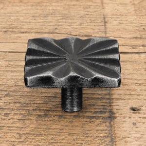 May include: A square cabinet knob with a dark, weathered metallic finish. The top surface features a radiating sunburst pattern of raised ridges and recessed grooves extending from the centre to the edges, supported by a cylindrical base.