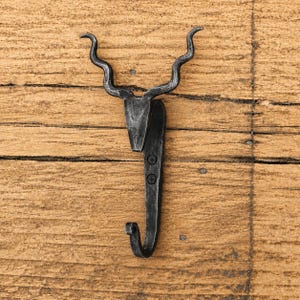 May include: A dark grey, hand-forged metal wall hook shaped like a stylised animal head with long, curved, wavy horns. The hook features a vertical mounting plate with two visible screw holes, extending downwards into a curved hook for hanging items.