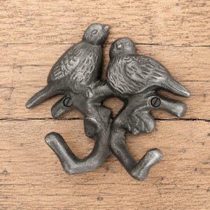 Könnte beinhalten: Ein dunkelgrauer Wandhaken aus Metall mit zwei Vögeln, die auf einem Ast sitzen. Das Design umfasst zwei geschwungene Haken, die vom unteren Teil des Astes ausgehen, sowie zwei sichtbare Schraublöcher zur Montage mit einer texturierten Gussmetalloberfläche.