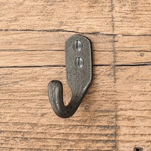 May include: A dark gray metal wall hook with a textured, weathered finish. The hook features a vertical mounting plate with two visible flat-head screws aligned vertically, and a single curved hook extending from the bottom of the plate.