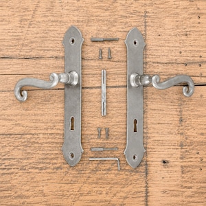 Op de afbeelding: Een set van twee zilverkleurige metalen deurklinken met lange rechthoekige schilden voorzien van sleutelgaten. De set bevat een centrale vierkante stift, vier kleine montageschroeven, twee draadstangen en een L-vormige inbussleutel.