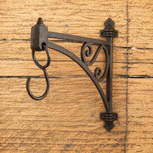 May include: A dark brown metal wall-mounted bracket featuring a vertical mounting plate with two screw holes, a horizontal support arm, and a decorative scrollwork design. A separate S-shaped hook hangs from a ring at the end of the arm.
