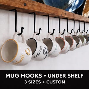 May include: A collection of white ceramic mugs with black rims suspended from black metal hooks beneath a wooden shelf. Each mug features a unique name or design, such as a horseshoe and a football. The text "MUG HOOKS • UNDER SHELF" is displayed below.
