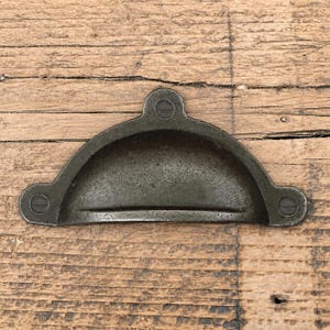 Peut inclure: Une poignée de tirage en métal gris foncé en forme de demi-cercle avec une zone de préhension encastrée. La quincaillerie présente trois points de fixation, chacun doté d'une vis à tête plate visible intégrée au design de la poignée.