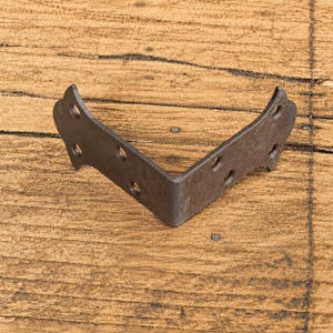 Può includere: Una staffa angolare in metallo marrone scuro con un profilo decorativo curvo. L'hardware a forma di L presenta sei fori di montaggio circolari preforati, con tre fori posizionati lungo ciascuno dei due bracci metallici piatti e perpendicolari.