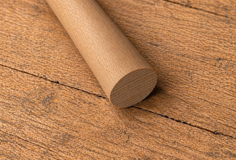 May include: A light brown, cylindrical wooden rolling pin with a rounded end, resting on a textured, brown wooden surface. The rolling pin is made of solid wood and has a smooth, natural finish. The wood grain is visible.