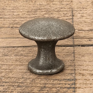 Puede incluir: Un pomo para mueble con forma de seta y un acabado metálico oscuro y desgastado. El pomo presenta una superficie superior ancha y redondeada y una base cilíndrica cónica que se ensancha en la parte inferior, con una textura irregular.