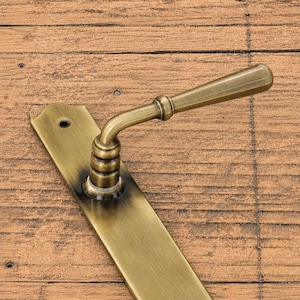 Puede incluir: Un juego de manija de puerta de latón con una placa trasera rectangular larga con un solo orificio para tornillo en la parte superior y una manija de palanca cilíndrica curva. El metal tiene un acabado antiguo cepillado con zonas sombreadas.