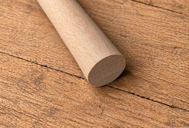 May include: A wooden rolling pin with a rounded end, resting on a wooden surface. The rolling pin is light brown and cylindrical, with visible wood grain. The background is a textured, warm-toned wooden surface.