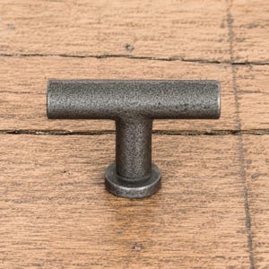 Peut inclure: Un bouton de meuble en forme de T avec une finition mate, texturée et gris foncé. La quincaillerie présente une barre cylindrique horizontale fixée à un montant vertical central relié à une base circulaire, formant une poignée fonctionnelle.