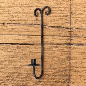 Puede incluir: Un candelabro de pared de metal negro con un vástago vertical largo y delgado que se curva en la parte inferior para sostener un plato circular y un soporte cilíndrico central, rematado con dos adornos de volutas simétricas.
