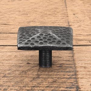 May include: A square cabinet knob with a dark grey, metallic finish and a hammered, dimpled texture across the top surface. The knob features a central post that connects to a flat, four-sided head with slightly bevelled edges and a dark patina.