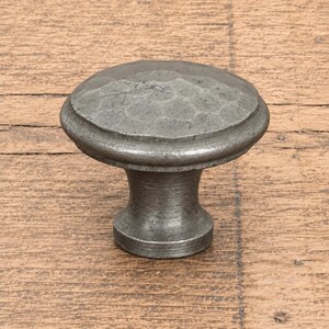Peut inclure: Un bouton de meuble rond avec une finition métallique gris foncé et une surface martelée et texturée sur le dessus. Le bouton présente une base lisse et effilée qui s'élargit vers le sommet circulaire, offrant une prise pour les tiroirs.
