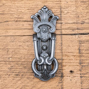 Op de afbeelding: Een donkergrijze metalen deurklopper met een langwerpig verticaal ontwerp, een decoratieve waaiervormige bovenkant, twee kleine bloemaccenten op de centrale plaat en een scharnierende U-vormige handgreep die uitloopt in een afgeronde basis.