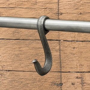 Puede incluir: Un gancho en forma de S de metal forjado gris oscuro colgado sobre una barra metálica horizontal de tono plateado. El gancho presenta un acabado mate y texturizado con un diseño curvo que rodea la parte superior de la barra y termina en punta.