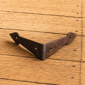 Pode incluir: Um suporte de canto metálico em forma de L, de cor castanho-escura, com um acabamento texturado e desgastado. O suporte apresenta um design pontiagudo em forma de seta em cada extremidade, com três orifícios circulares pré-perfurados.