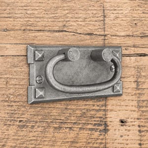 Op de afbeelding: Een rechthoekige metalen ladehandgreep met een donkergrijze afwerking en een getextureerd oppervlak. Het beslag is voorzien van een centrale valgreep, vier piramidevormige hoekaccenten en twee zichtbare platte schroeven voor bevestiging.