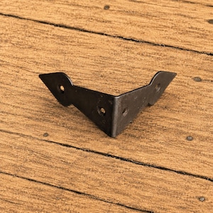 Könnte beinhalten: Ein dunkelgrauer Eckwinkel aus Metall mit V-förmigem Design. Der Winkel verfügt über drei kreisförmige Schraublöcher, eines auf jeder Seite und eines am Mittelpunkt, mit spitz zulaufenden Enden an den beiden Armen des Metallwinkels.