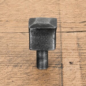 Könnte beinhalten: Ein dunkelgrauer, quadratischer Möbelknopf aus Metall mit einer texturierten, gehämmerten Oberfläche. Der Knopf verfügt über eine leicht erhöhte, abgeschrägte Oberseite und einen zylindrischen Sockel, der nach unten verläuft und Halt bietet.