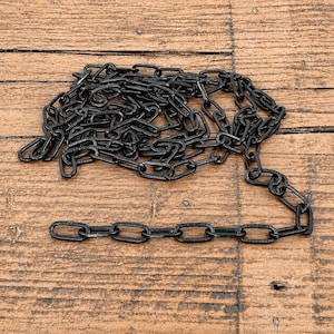 Op de afbeelding: Een matzwarte metalen ketting met langwerpige, ovale schakels. De ketting is gerangschikt in een losse, opgerolde hoop met een enkel recht segment dat uit het hoofdcluster steekt, wat de uniforme vorm van de schakels goed laat zien.