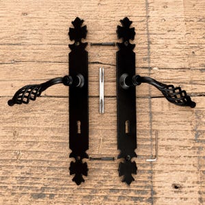 May include: A set of two matte black door handles with long rectangular backplates featuring decorative finials at the top and bottom. The handles have a cage-like twisted design. Included are two black screws, a metal spindle, and an L-shaped hex key.