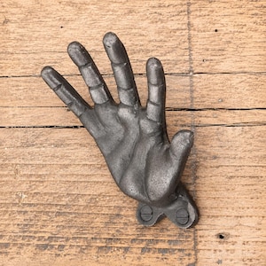 May include: A dark gray, textured metal wall hook shaped like an open human hand with fingers extended upward. The base of the hand features two circular mounting points with visible screws for attachment to a flat surface, showcasing a rugged finish.