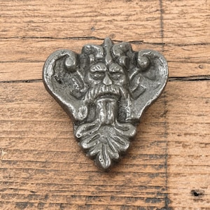Könnte beinhalten: Ein dekoratives Objekt in Silberoptik mit einem skulptierten Gesicht, das einen markanten Schnurrbart und Bart aufweist, eingebettet in ein symmetrisches Blattdesign mit geschwungenen Ornamenten, die von der zentralen Figur ausgehen.