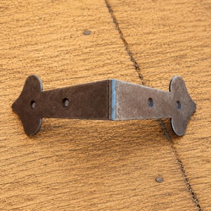 Può includere: Una staffa angolare in metallo grigio scuro con un'estremità appuntita a forma di picca su ciascun lato. La staffa presenta un design piegato a forma di L con due fori circolari per viti visibili su ciascuno dei due bracci piatti di rinforzo.