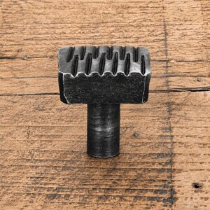 May include: A dark grey metal cabinet knob featuring a rectangular head with a series of vertical grooves along the top edge and a cylindrical base. The hardware has a textured, matte finish with visible signs of metalwork and a solid construction.