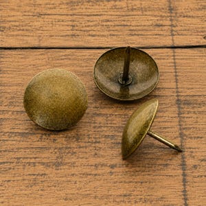 May include: Three antique brass upholstery tacks with a smooth, domed head and a sharp, pointed metal nail extending from the center of the underside. The finish has a weathered, metallic texture with subtle variations in color and tone.