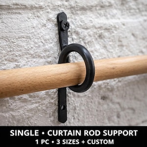 Puede incluir: Un soporte de barra de cortina negro, individual, montado en una pared de ladrillo blanco. El soporte presenta un anillo circular y un soporte vertical, que sujeta una barra de madera. El texto en la parte inferior dice "SINGLE • CURTAIN ROD SUPPORT".