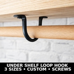 Könnte beinhalten: Schwarze Metall-Haken unter dem Regal, die an einem Holzregal befestigt sind und eine Holzstange halten. Die Haken sind gebogen und zum Aufhängen von Gegenständen konzipiert. Der Text unten lautet: UNDER SHELF LOOP HOOK, 3 SIZES, CUSTOM, SCREWS.