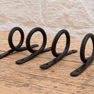 May include: Four black metal wall hooks, each featuring a circular ring attached to a flat, elongated base plate with two pre-drilled screw holes for mounting. The hooks have a dark, matte finish and a simple, functional design for hanging items.