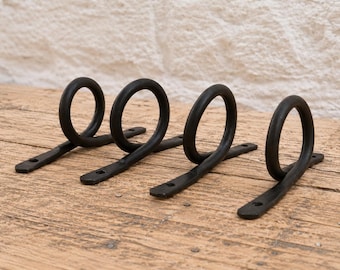 May include: Four black metal wall hooks, each featuring a circular ring attached to a flat, elongated base plate with two pre-drilled screw holes for mounting. The hooks have a dark, matte finish and a simple, functional design for hanging items.