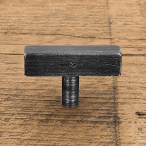 May include: A T-shaped cabinet knob with a dark, weathered charcoal finish. The horizontal bar features a textured, brushed metal surface and a small, stamped four-petal flower detail centred on the front face, supported by a cylindrical base.