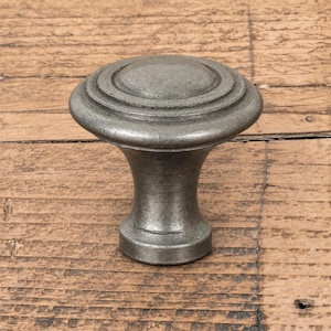 Puede incluir: Un pomo de mueble redondo con un acabado metálico gris oscuro desgastado. El pomo presenta una superficie superior lisa y ligeramente abombada con crestas circulares concéntricas en el borde, estrechándose hacia una base cilíndrica plana.