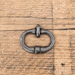 May include: A dark gray metal buckle with an oval frame and a central vertical bar. The buckle has a textured, weathered finish with visible signs of wear, featuring a smooth, rounded shape that connects at the top and bottom of the oval frame.